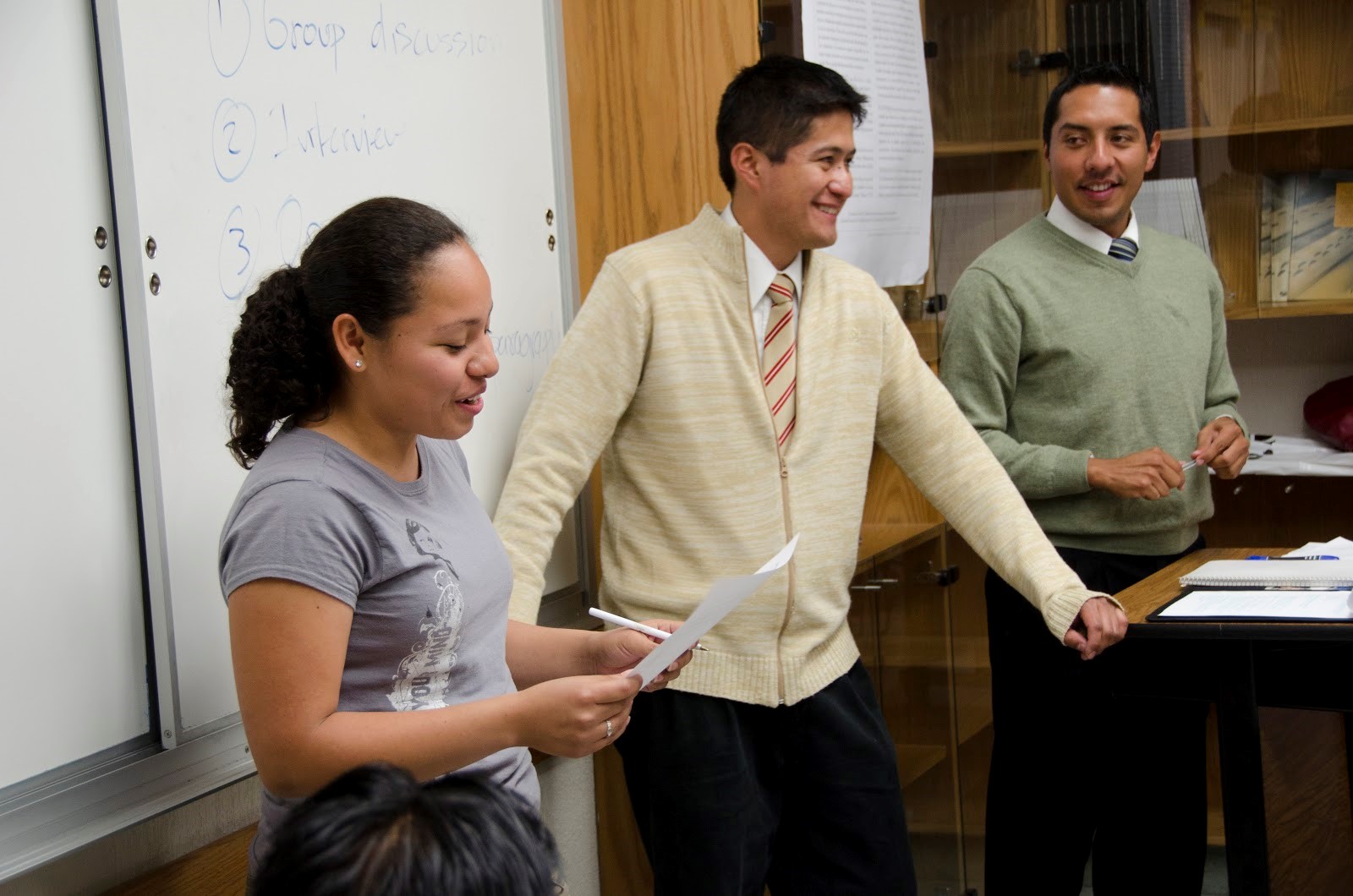 A woman presents in front of a classroom alongside two men.