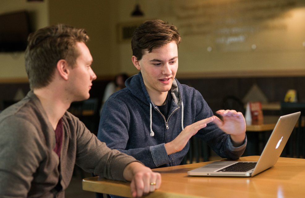 One students explains something to another student while looking at a computer.