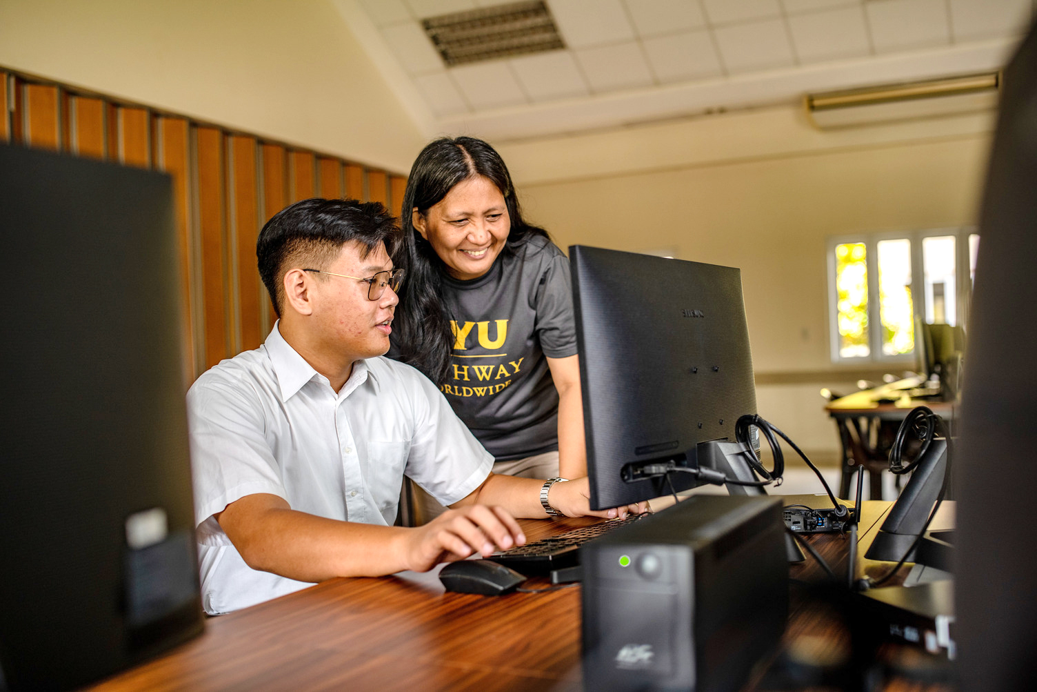 Two BYU-Pathway students in Cebu City, Philippines collaborate at a desktop computer, smiling as they work together in a classroom setting.