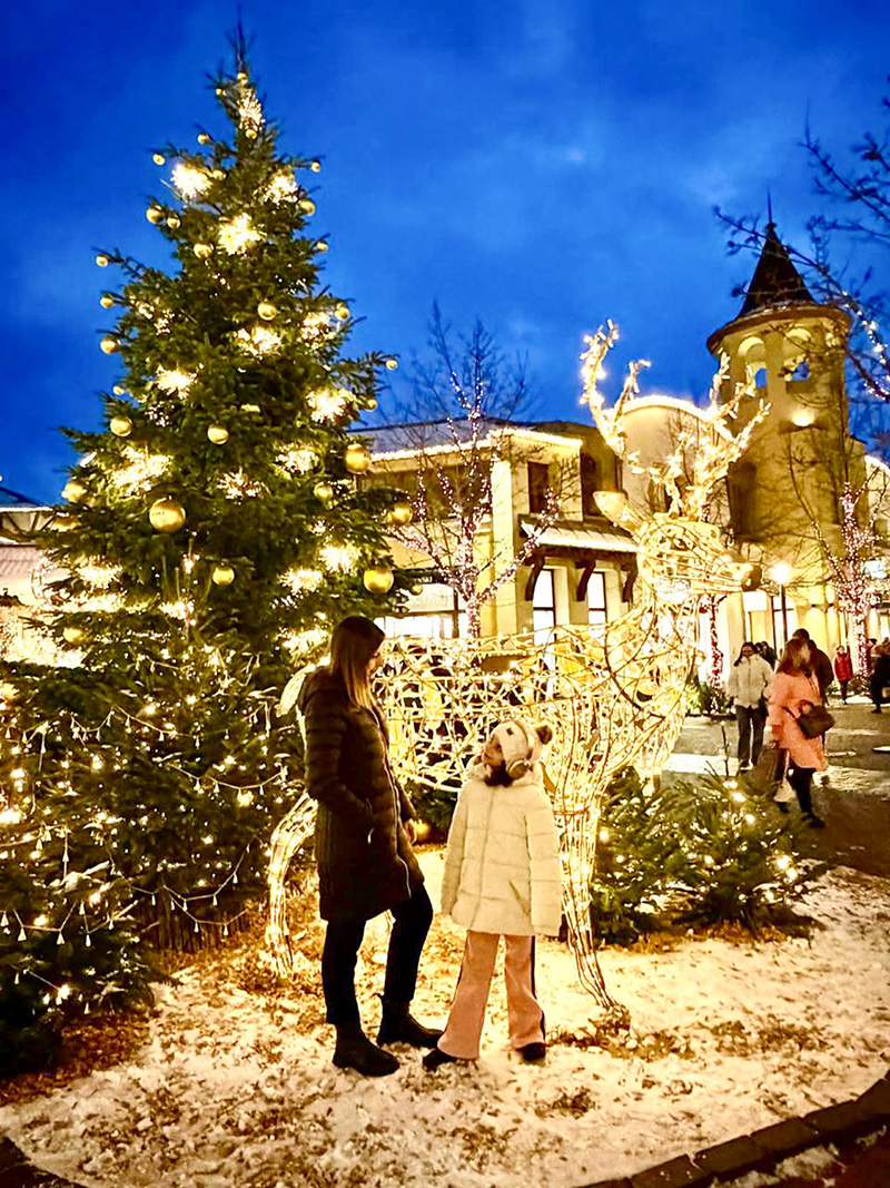 BYU-Pathway student Alina and her daughter stand beside a lit reindeer sculpture and decorated Christmas tree in a festive outdoor setting at dusk, surrounded by holiday lights and light snow.