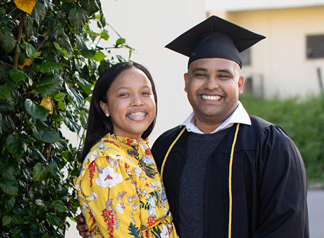 BYU-Pathway student Juan-Pablo stands in cap and gown beside his wife, smiling proudly outdoors near green foliage.