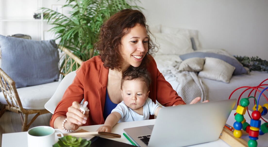 Attractive young dark skinned woman working at desk at home using laptop, holding baby on her lap. Portrait of smiling mother writing post on moms blog while her infant son playing with toy