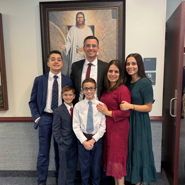 The Maia family, including Karine, her husband, and their four children, stand together in front of a picture of Christ with their arms around one another.