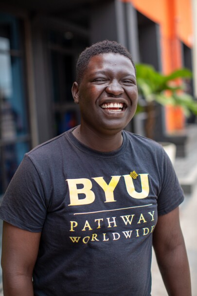 Smiling BYU-Pathway student wearing a BYU-Pathway Worldwide t-shirt stands outdoors.