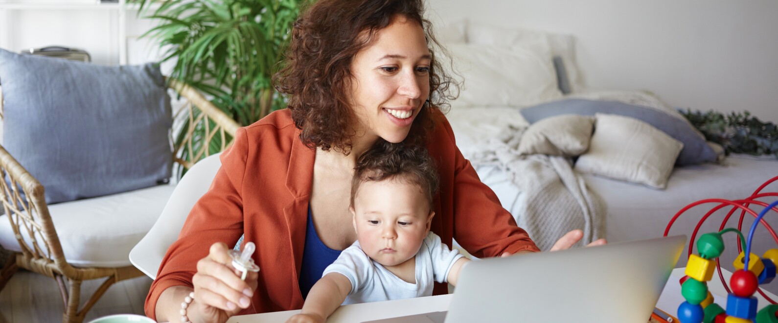 Attractive young dark skinned woman working at desk at home using laptop, holding baby on her lap. Portrait of smiling mother writing post on moms blog while her infant son playing with toy