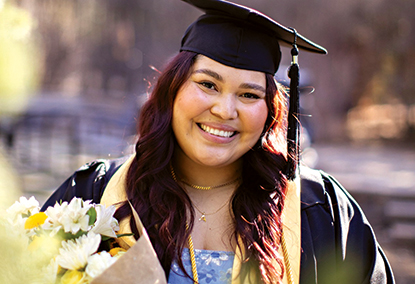 BYU-Pathway graduate Ale Angula, originally from Costa Rica, smiles in cap and gown while holding a bouquet of flowers outdoors.