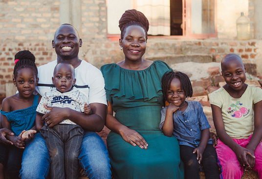 BYU-Pathway student Faith Ssenyonga from Uganda sits with her family outside their home, smiling together for a family portrait.