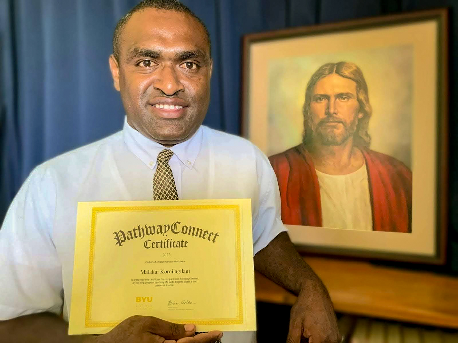 Malakai Koroilagilagi, a BYU-Pathway student from Fiji, holds his PathwayConnect certificate while standing beside a framed portrait of Jesus Christ, smiling proudly at the camera.